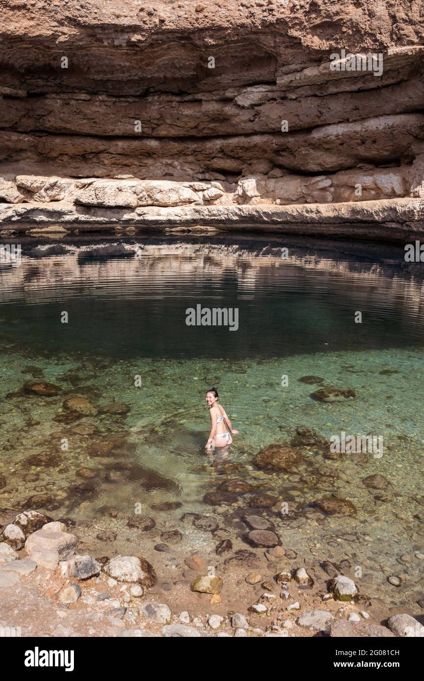 Femme asiatique détendue regardant de nouveau à l'appareil photo sur l'eau transparente de Bimmah Sinkhole entouré de rochers rugueux pendant le voyage à Oman Banque D'Images