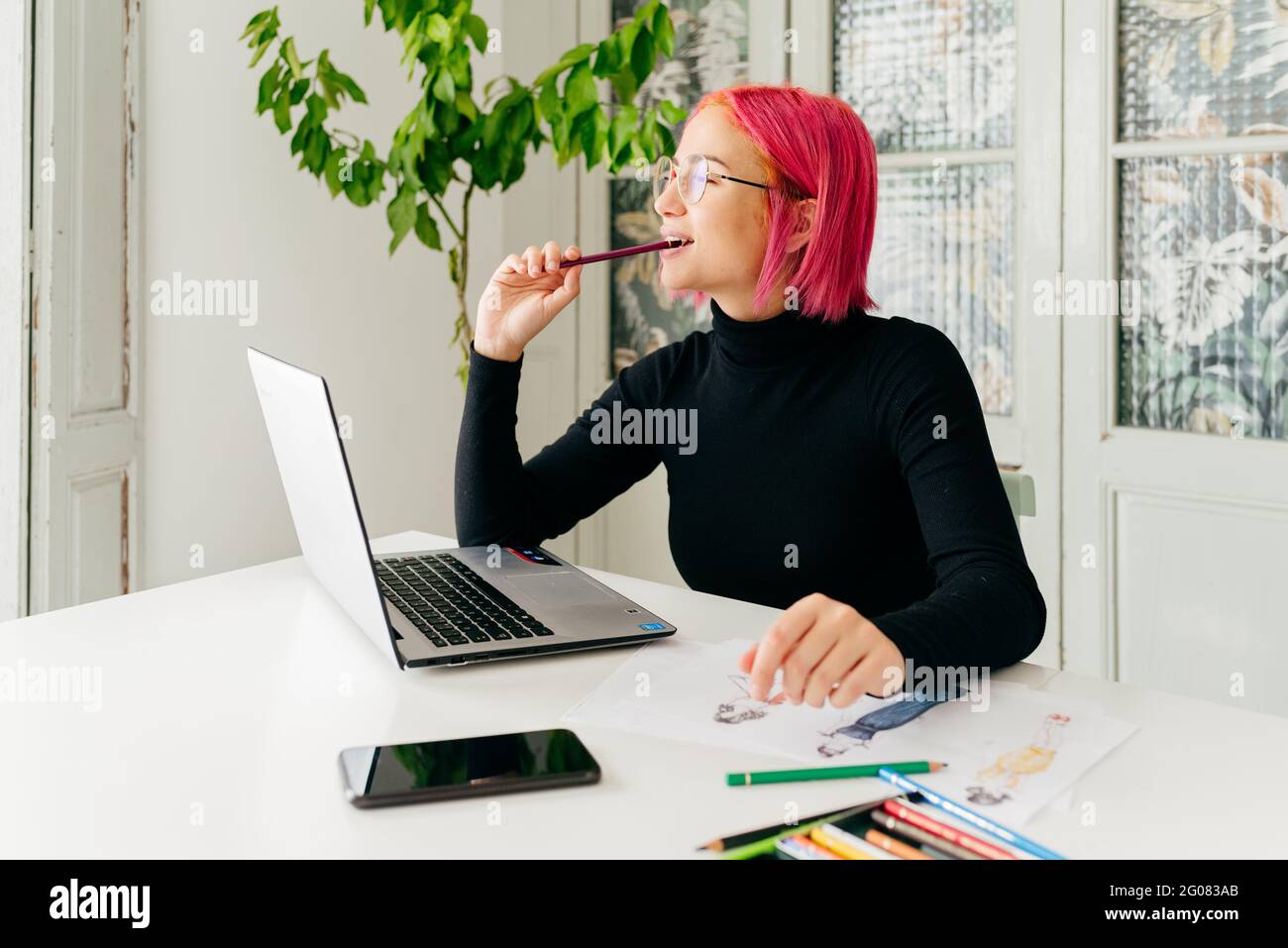 Femme ingénieuses designer freelance tenue décontractée et lunettes de travail à la table avec ordinateur portable et dessin de croquis de mode avec des crayons Banque D'Images