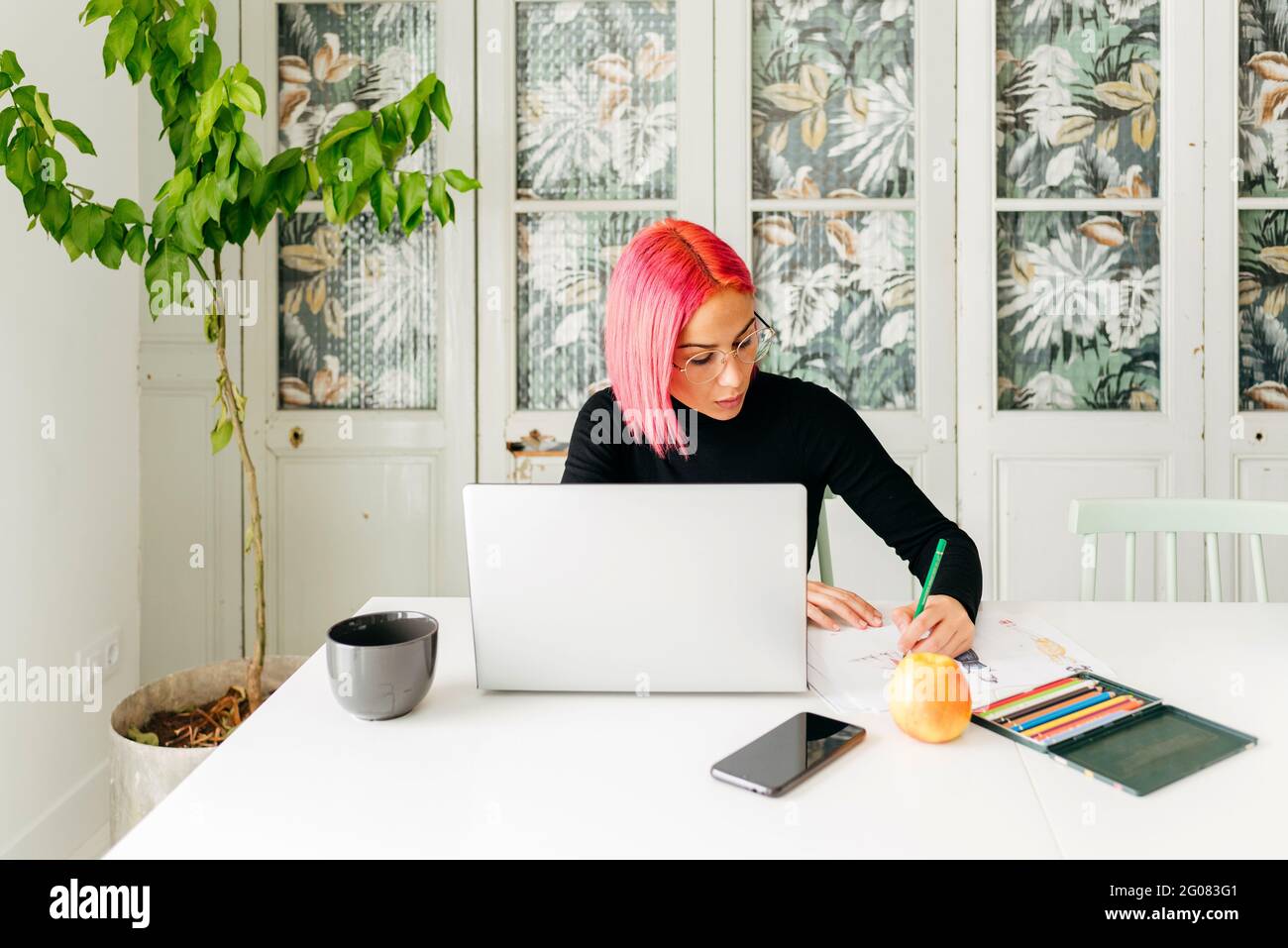 Femme ingénieuses designer freelance tenue décontractée et lunettes de travail à la table avec ordinateur portable et dessin de croquis de mode avec des crayons Banque D'Images