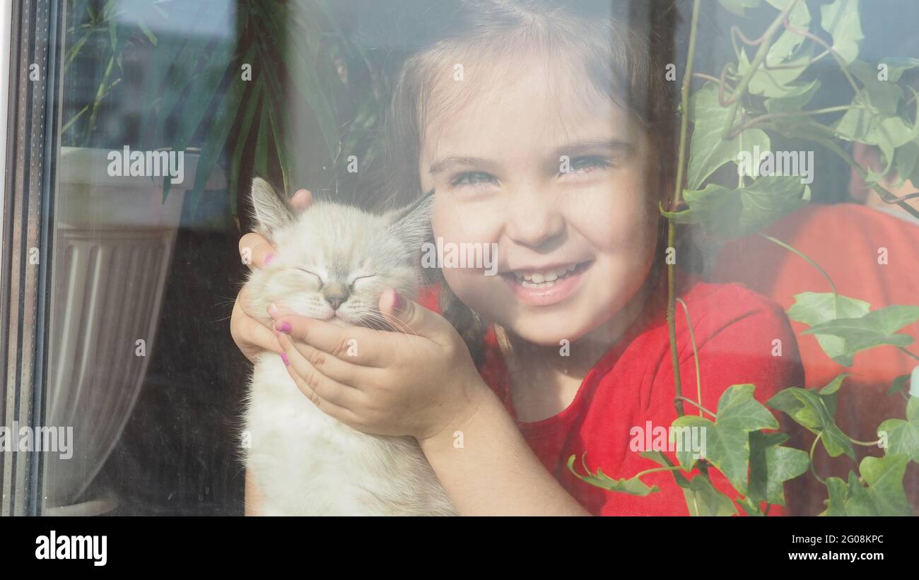 Belle petite fille souriant et regardant par la fenêtre. Un enfant regarde la fenêtre. Portrait d'un gamin joyeux. Auto-distanciation à la maison pendant Coro Banque D'Images