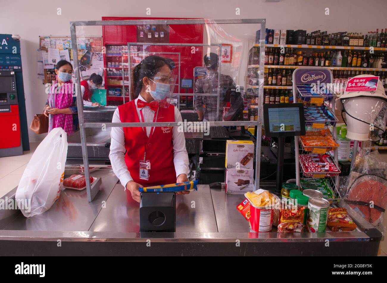 En raison d'une épidémie de COVID - 19, un caissier de supermarché, au comptoir des caisses, porte un masque facial et un écran facial de protection tout en se tenant derrière un écran en plastique fait maison pendant la pandémie du coronavirus. Phnom Penh, Cambodge. 19 décembre 2020. © Kraig Lieb Banque D'Images