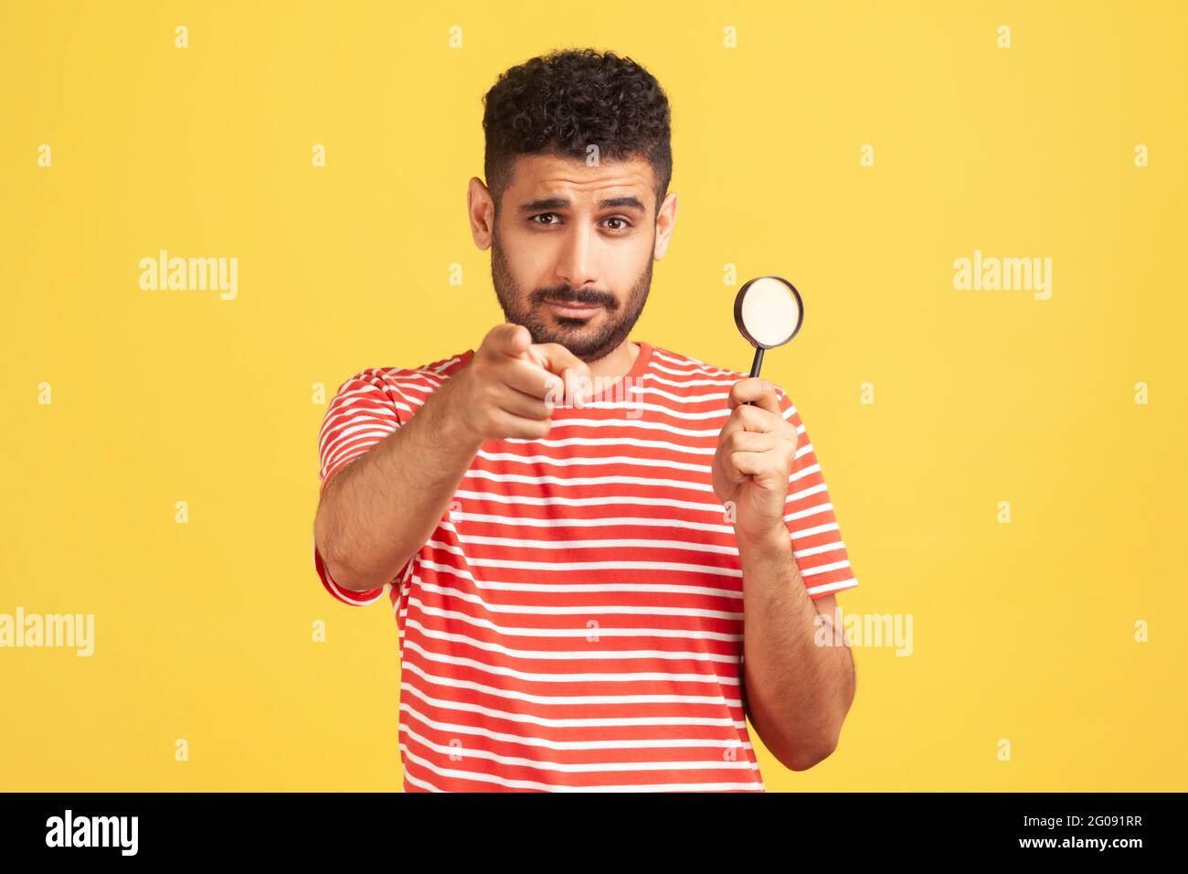 Homme sérieux confiant détective privé dans un t-shirt rayé tenant dans la main loupe et le doigt pointant sur vous regardant la caméra, l'espionnage. Indo Banque D'Images