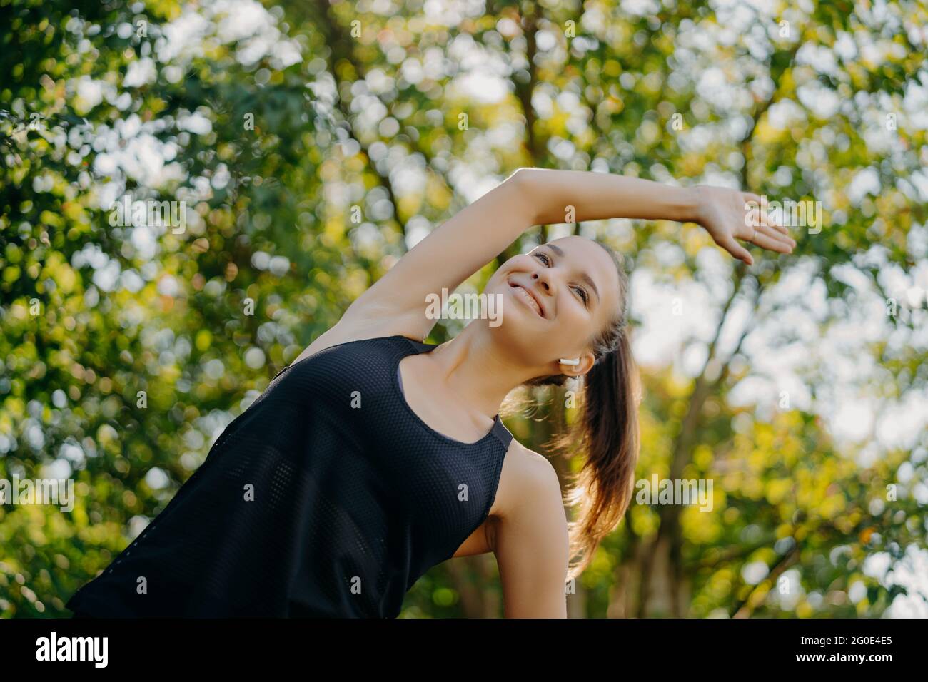 La femme sportive se réchauffe et les exercices d'étirement s'étire de côté a contenu l'expression du visage vêtu de vêtements actifs pose sur le fond de la nature Banque D'Images