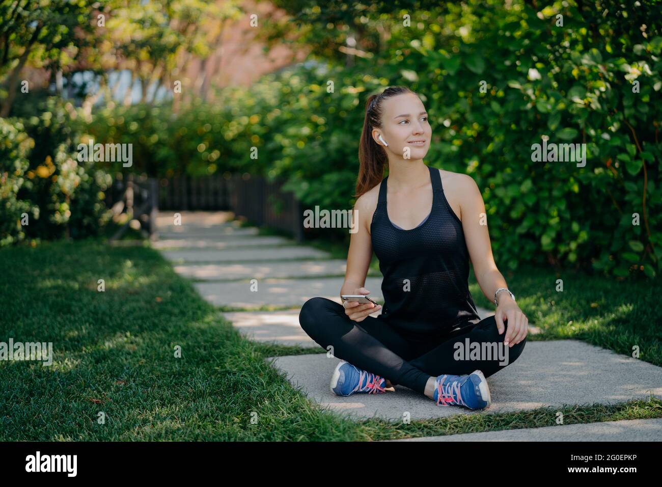 Une jeune femme est assise croise les jambes regarde de côté avec expression satisfait utilise smartphone a pause après l'entraînement cardio habillé en vêtements actifs et sneaker Banque D'Images