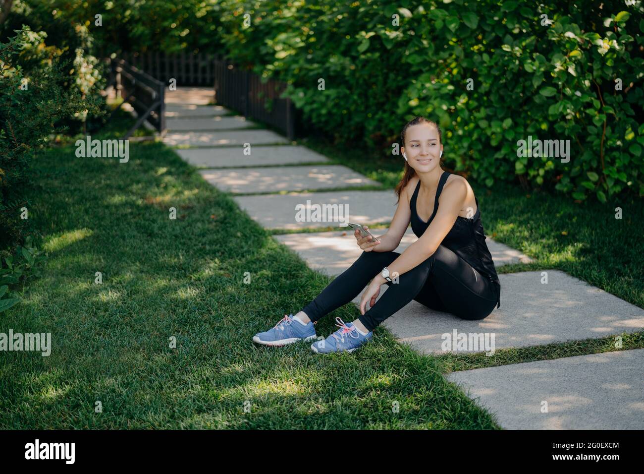 Active brunette European Woman télécharge de la musique sur smartphone pour l'entraînement actif dehors a pause après l'entraînement vêtu de vêtements actifs partage du contenu Banque D'Images