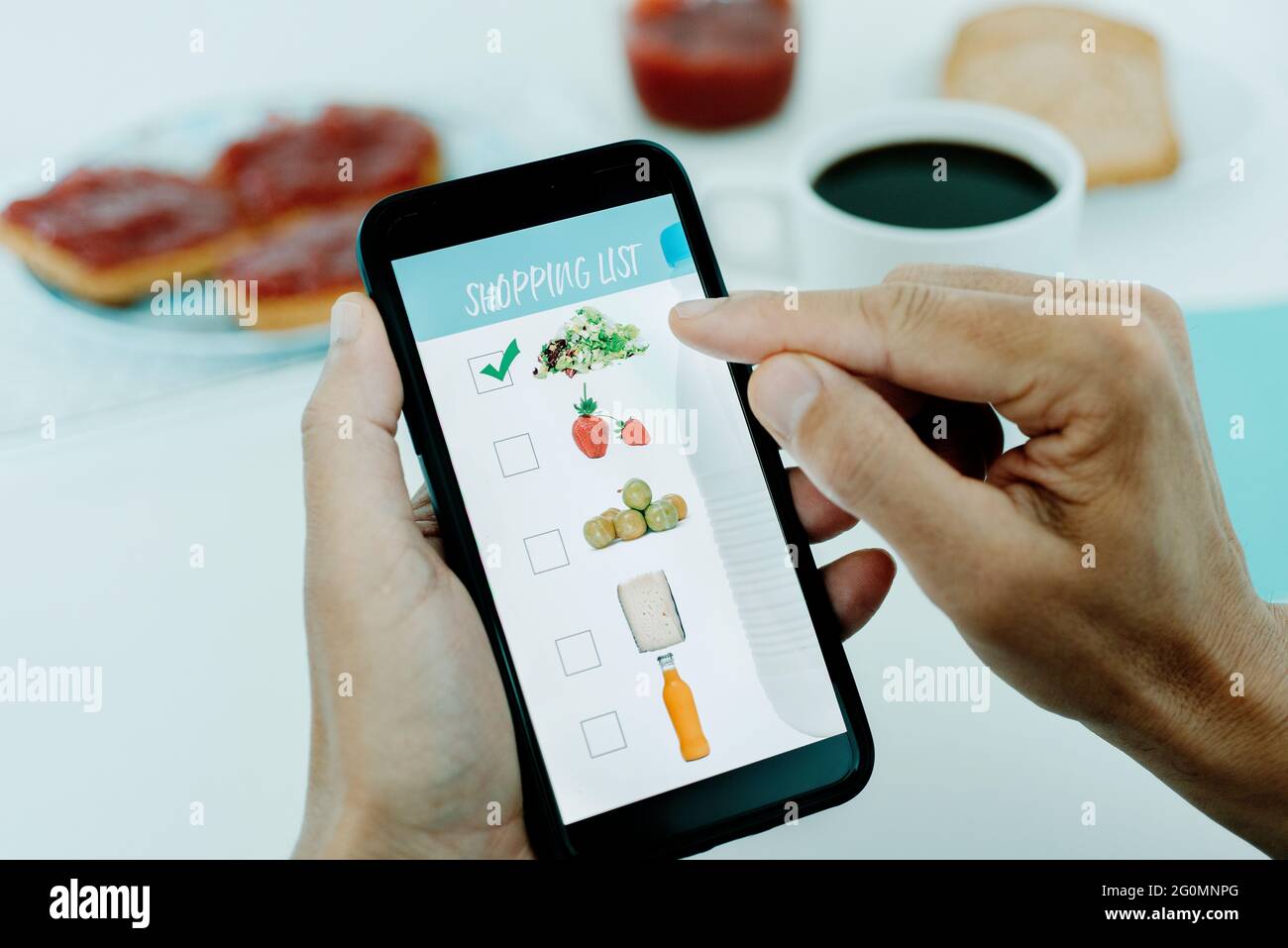 gros plan d'un jeune homme caucasien, assis à une table pour le petit déjeuner, faisant sa liste d'achats dans son smartphone, avec des photos de différents produits ma Banque D'Images
