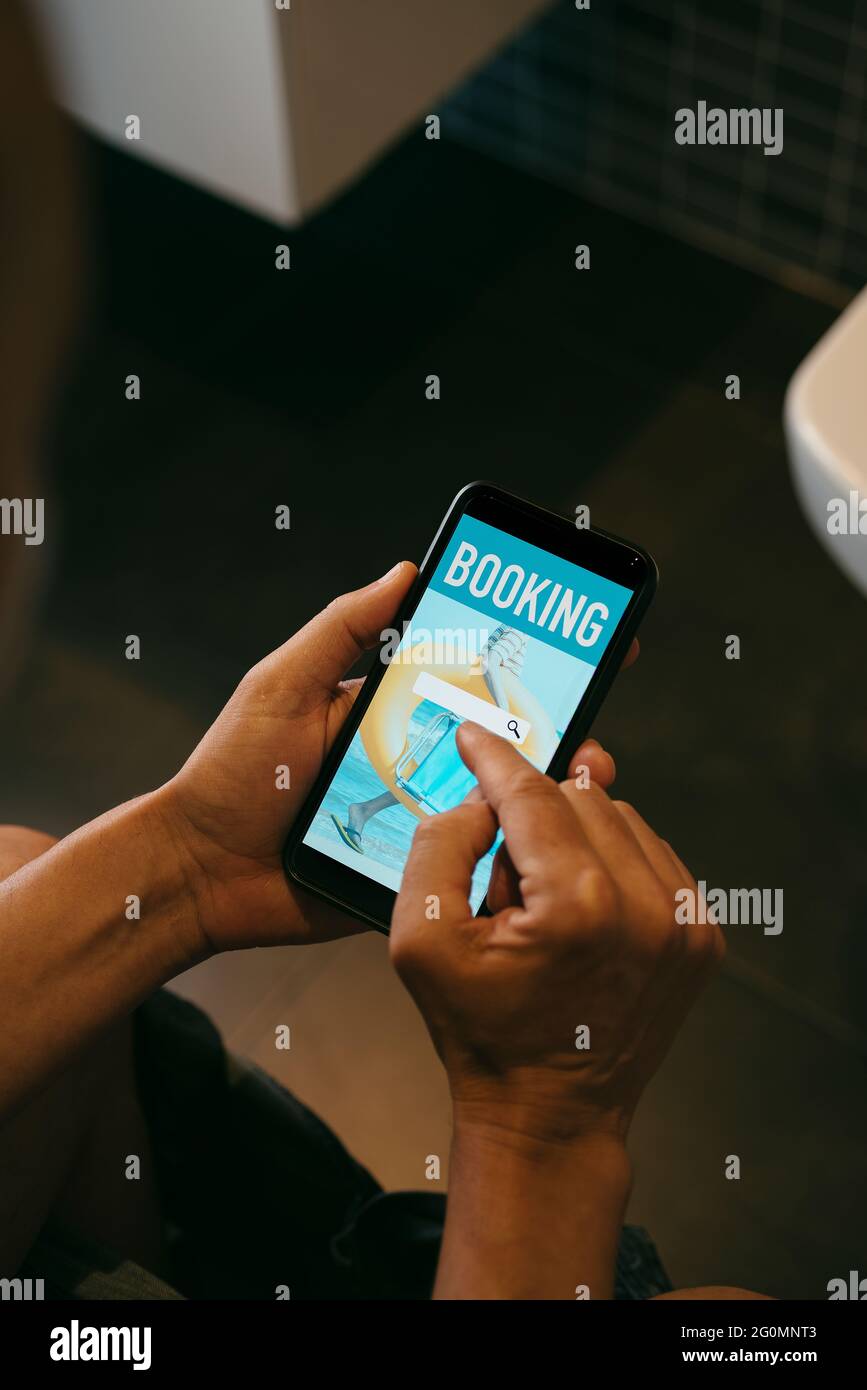 gros plan d'un jeune homme caucasien, assis dans les toilettes, réservant ses vacances en ligne avec son smartphone, avec un moteur de recherche simulé dans son écran Banque D'Images