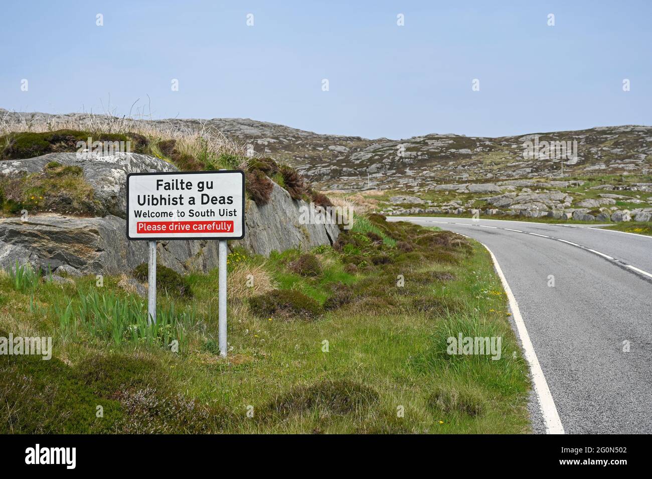 Bienvenue à South Uist signe en anglais et gaélique écossais. Arrière-plan de la route, de l'herbe et du paysage rocheux. Jour ensoleillé avec ciel bleu, pas de personnes. Banque D'Images