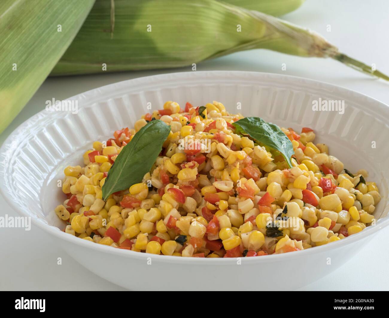Un bol de service blanc contenant un plat d'accompagnement de maïs et de poivron rouge coloré se trouve sur une table avec une pile d'épis de maïs non décortiqués fraîchement cueillis dans le bac Banque D'Images