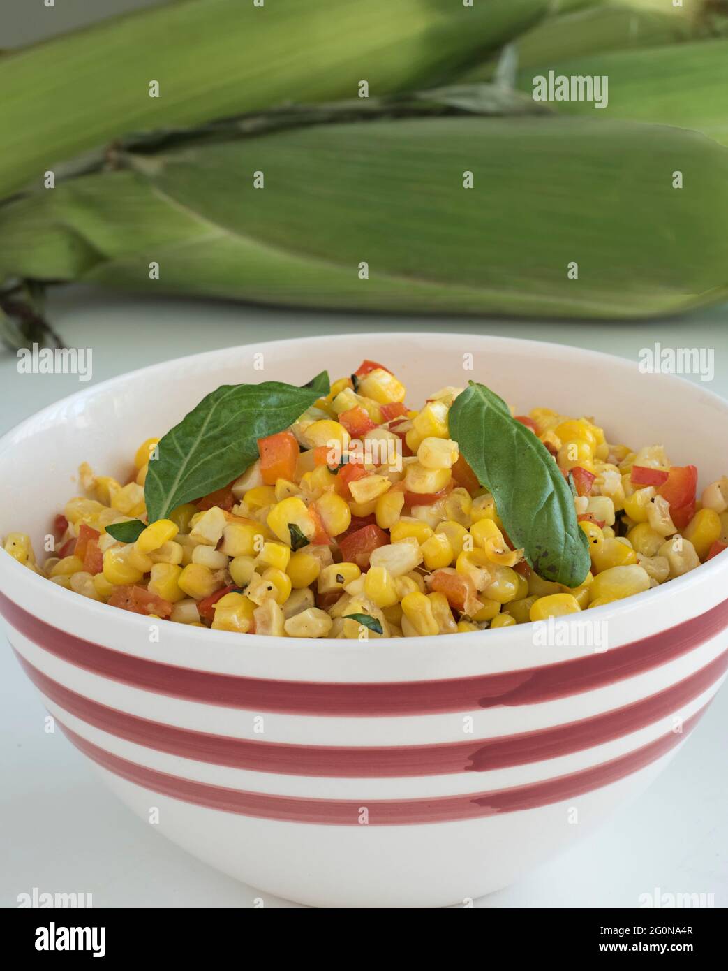 Un bol de service à rayures rouges et blanches rempli d'une casserole de maïs et de poivre rouge se trouve sur une table devant une pile de maïs frais non décortiqué Banque D'Images