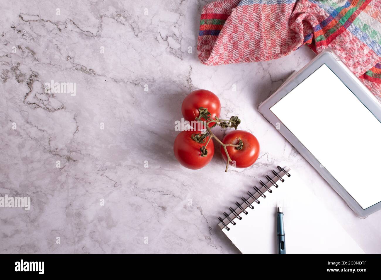 Trois tomates avec des branches sur une table en marbre accompagnées d'un carnet, d'une tablette et d'une torchon. Banque D'Images