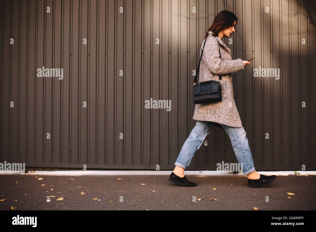 Vue latérale d'une femme non binaire utilisant un smartphone qui marche contre le mur Banque D'Images