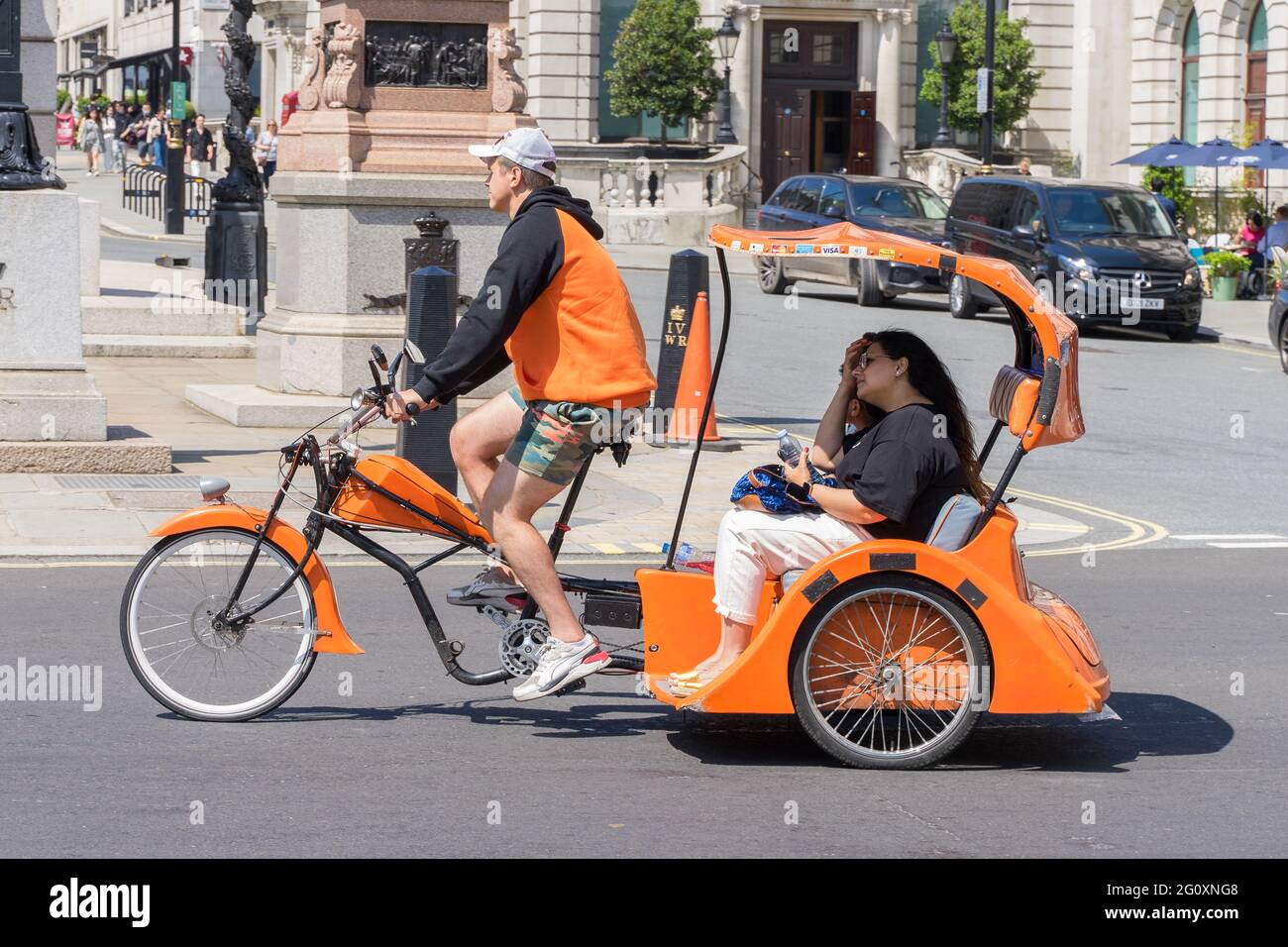 Un pousse-pousse orange avec des touristes à l'arrière dans les rues de Londres lors d'une chaude journée d'été. Banque D'Images