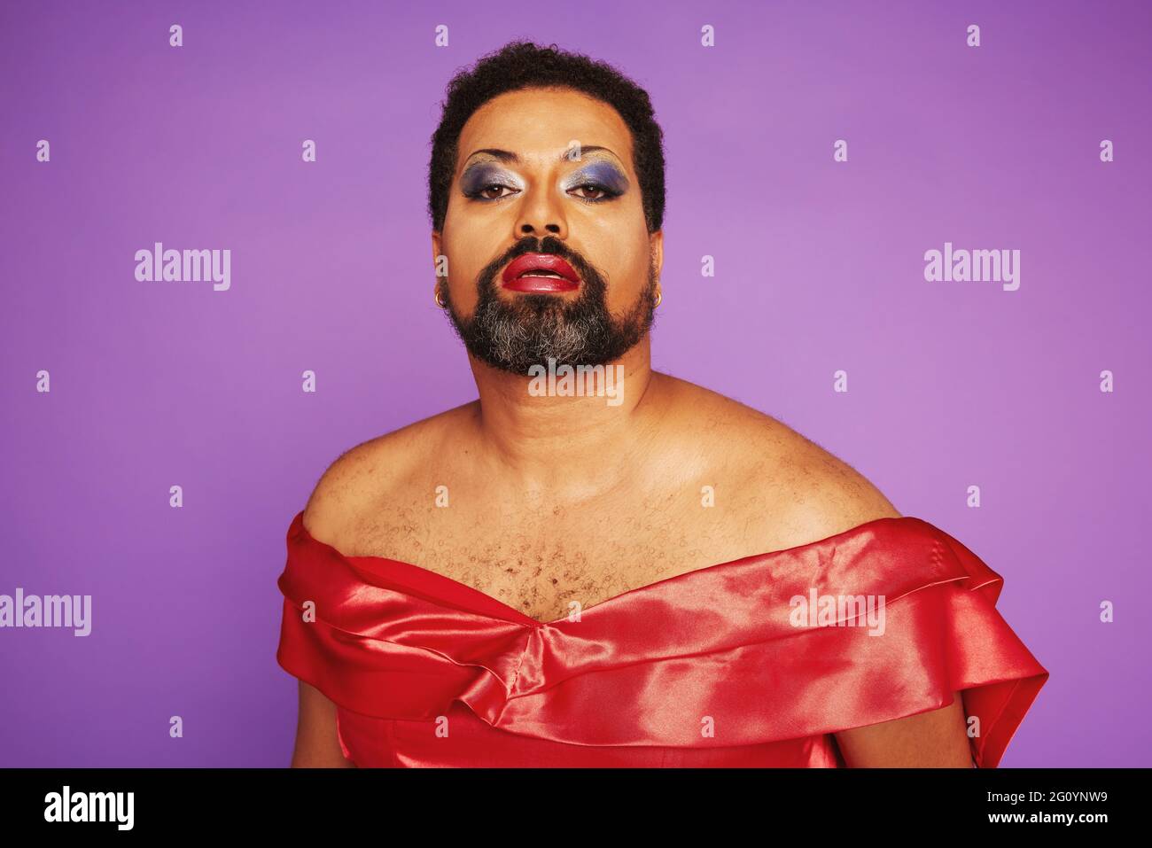 Portrait d'un homme avec une barbe portant une robe rouge et un maquillage féminin. Élégante reine de drag sur fond violet. Banque D'Images