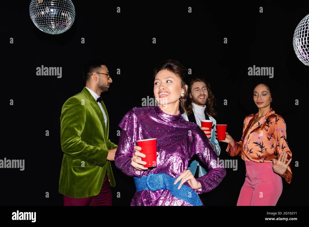 jeune adulte espiègle femme asiatique debout à la main sur la posture de la hanche près des amis interraciaux dansant sur fond noir Banque D'Images