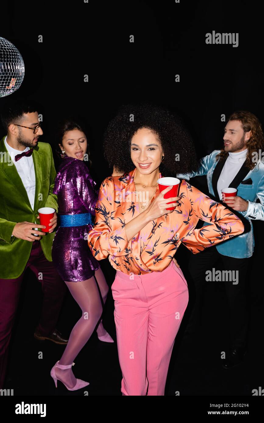 jeune femme afro-américaine adulte à la main sur la posture de la hanche avec des amis multiraciaux flous dansant sur fond noir Banque D'Images