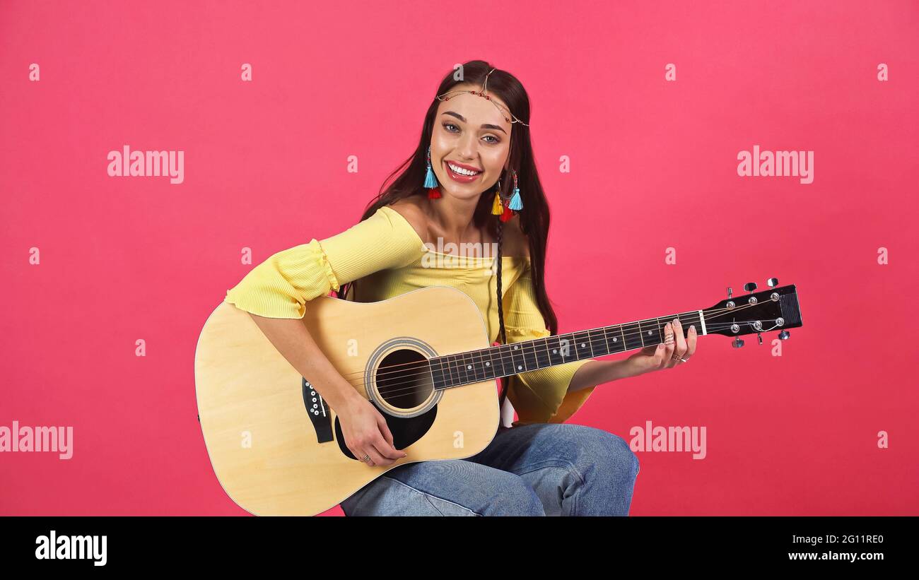 bonne jeune femme adulte en boucles d'oreilles colorées et accessoire de  cheveux assis avec guitare isolée sur rose Photo Stock - Alamy