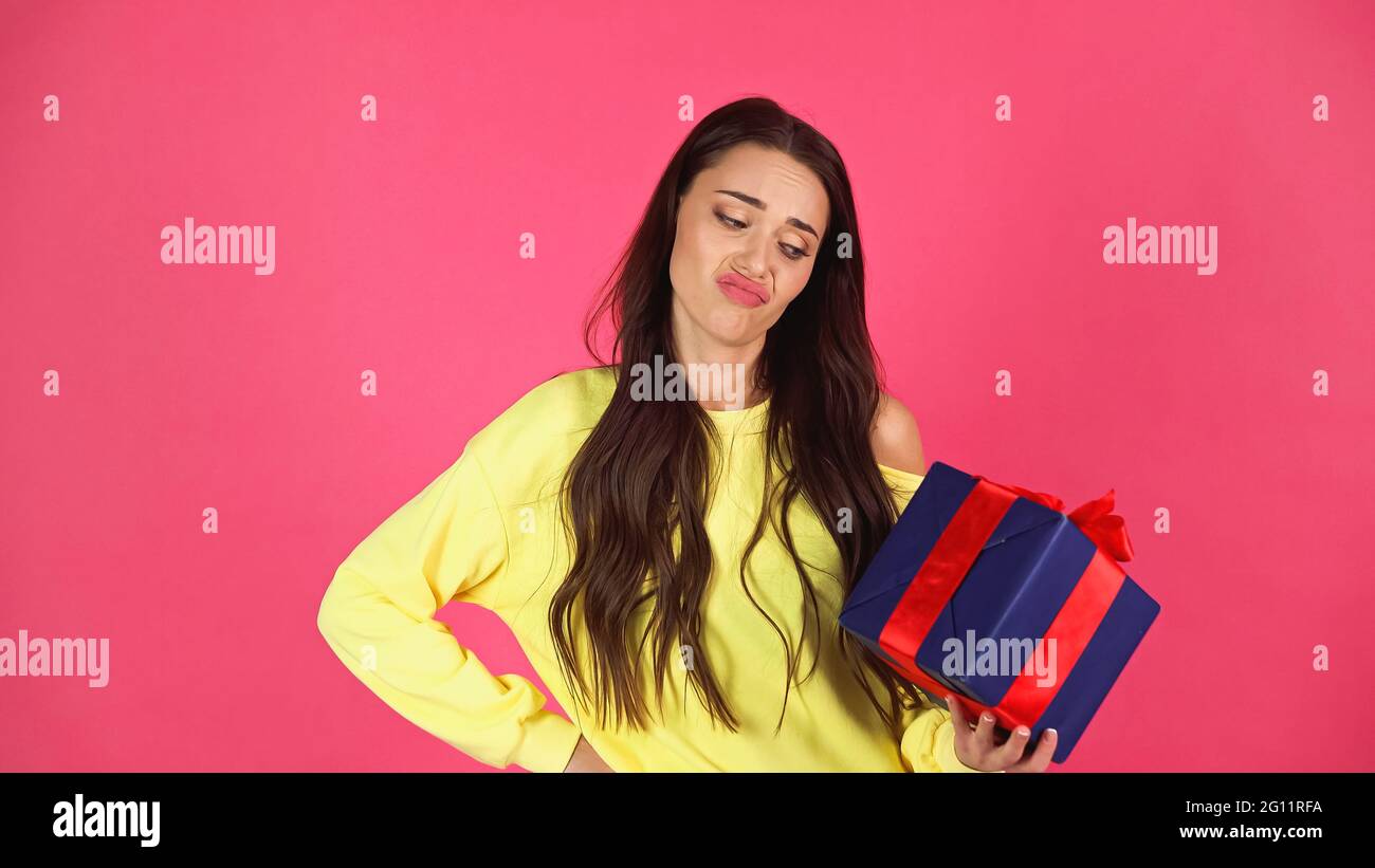 jeune femme insatisfaite adulte tenant une boîte cadeau avec la main sur la hanche pose isolée sur rose Banque D'Images