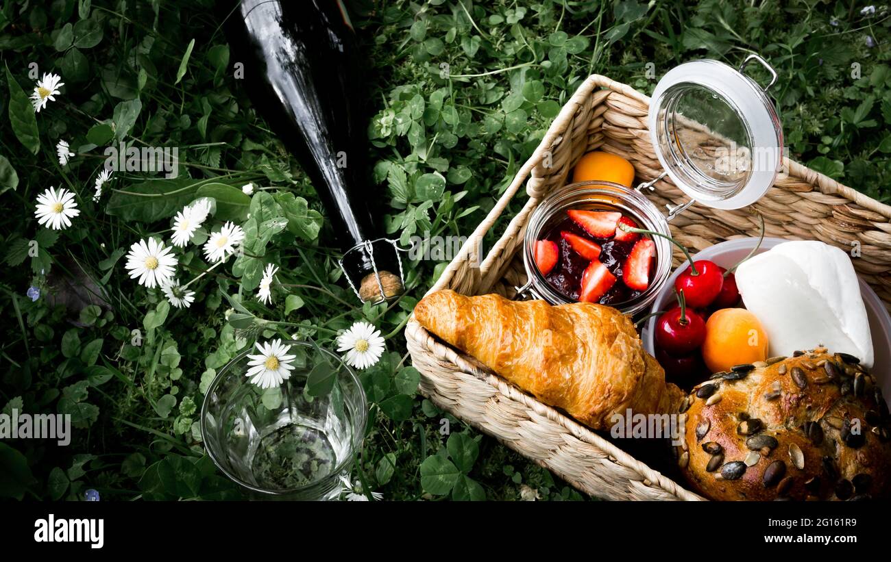 Panier pique-nique avec croissant français, fromage, confiture de fraises, cerises, abricots et pain de seigle avec graines de citrouille et bouteille de cidre sur prairie. Banque D'Images