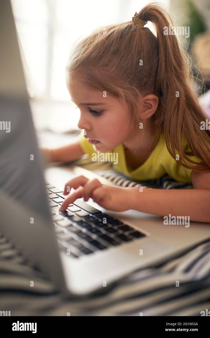 Une petite fille tapant sur l'ordinateur portable sur le lit dans une atmosphère détendue à la maison. Jouer, enfance, maison Banque D'Images