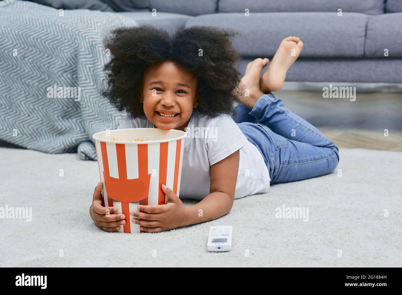 Petite fille afro-américaine tient un grand seau à pop-corn, couché à l'étage de la maison tout en regardant un film de dessin animé au cinéma à domicile Banque D'Images