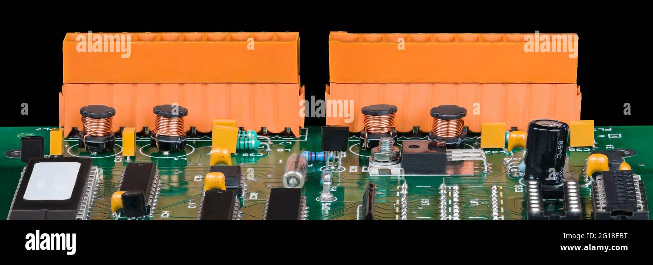 Détail panoramique des composants électroniques sur une carte de circuit imprimé verte. Puces, bobines cylindriques, transistors, condensateurs et deux borniers orange. Banque D'Images