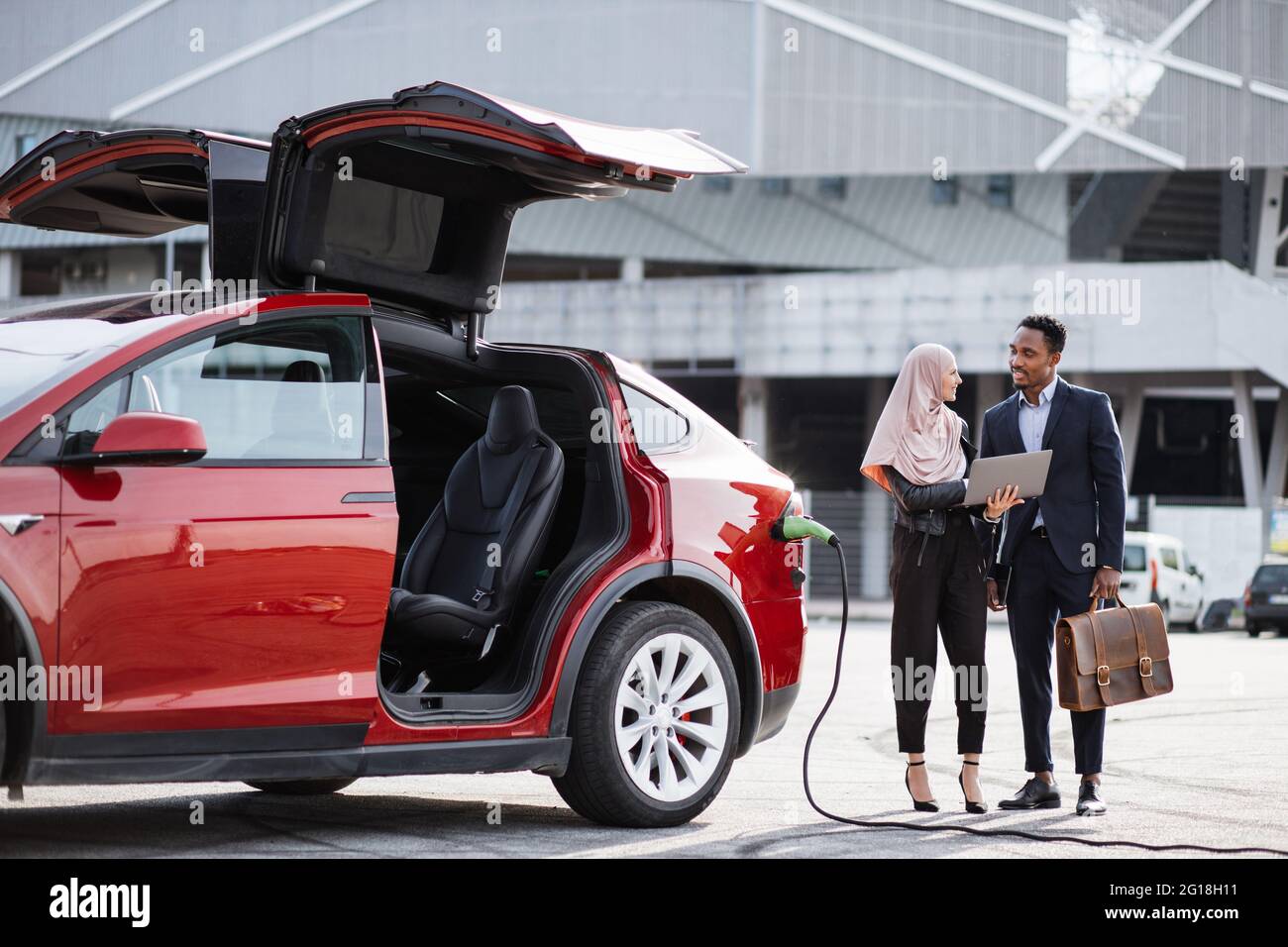 La voiture électrique rouge se recharge alors que la femme musulmane et l'homme africain parlent à proximité. Vendeur féminin dans hijab utilisant un ordinateur portable tout en faisant la démonstration de l'automobile moderne à un client masculin en costume. Banque D'Images