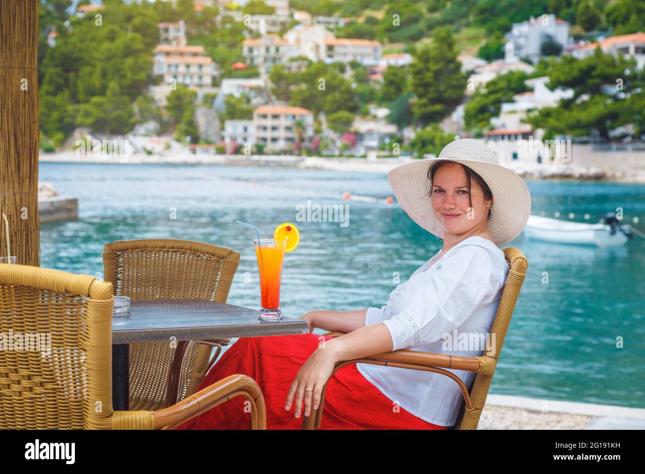 Une jolie femme heureuse se détendant et buvant un cocktail au bar de la plage en plein air. Belle femme portant une jupe rouge, une chemise blanche et un chapeau de paille à la plage Banque D'Images