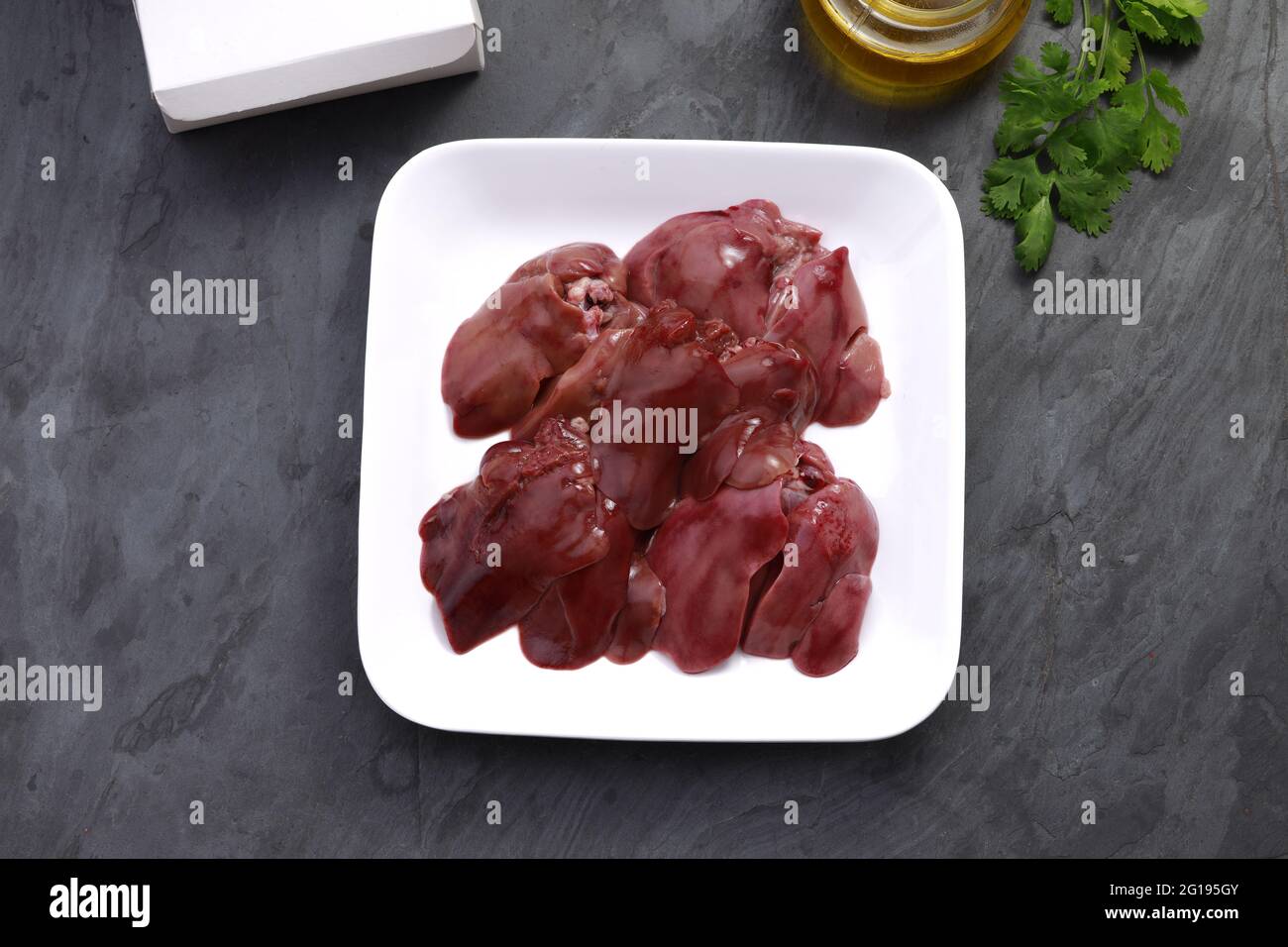 Foie de poulet cru magnifiquement arrangé dans une plaque de céramique blanche avec feuilles de coriandre, huile de cuisson et boîte d'emballage sur fond et placé sur le t gris Banque D'Images