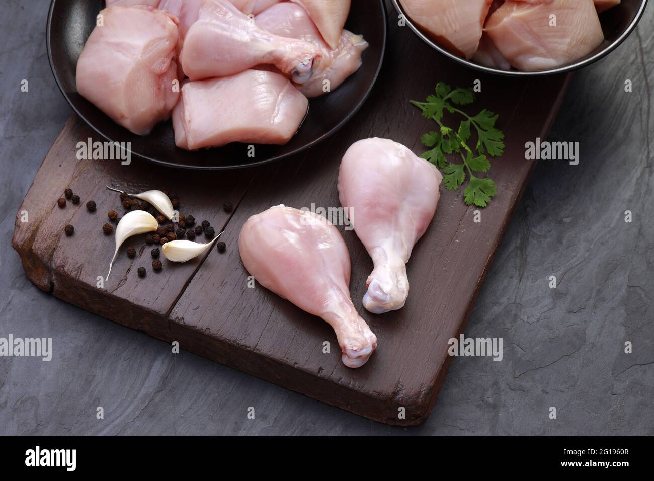 Ensemble de coupes de poulet cru, combinaison de coupes de curry de poulet sans peau, de coupes de poulet et de deux pilons disposés sur une planche en bois avec des feuilles de coriandre Banque D'Images