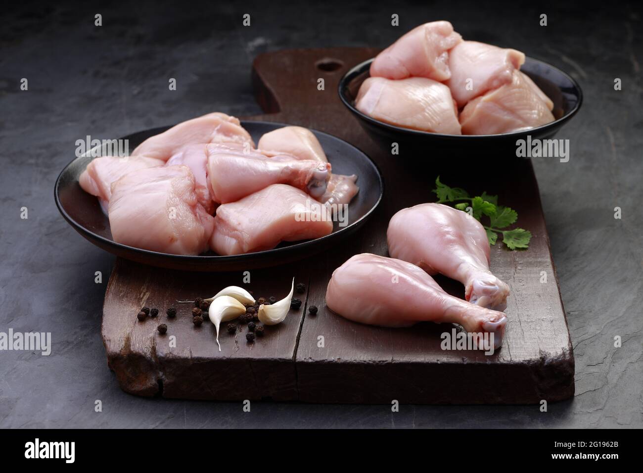 Ensemble de coupes de poulet cru, combinaison de coupes de curry de poulet sans peau, de coupes de poulet et de deux pilons disposés sur une planche en bois avec des feuilles de coriandre Banque D'Images