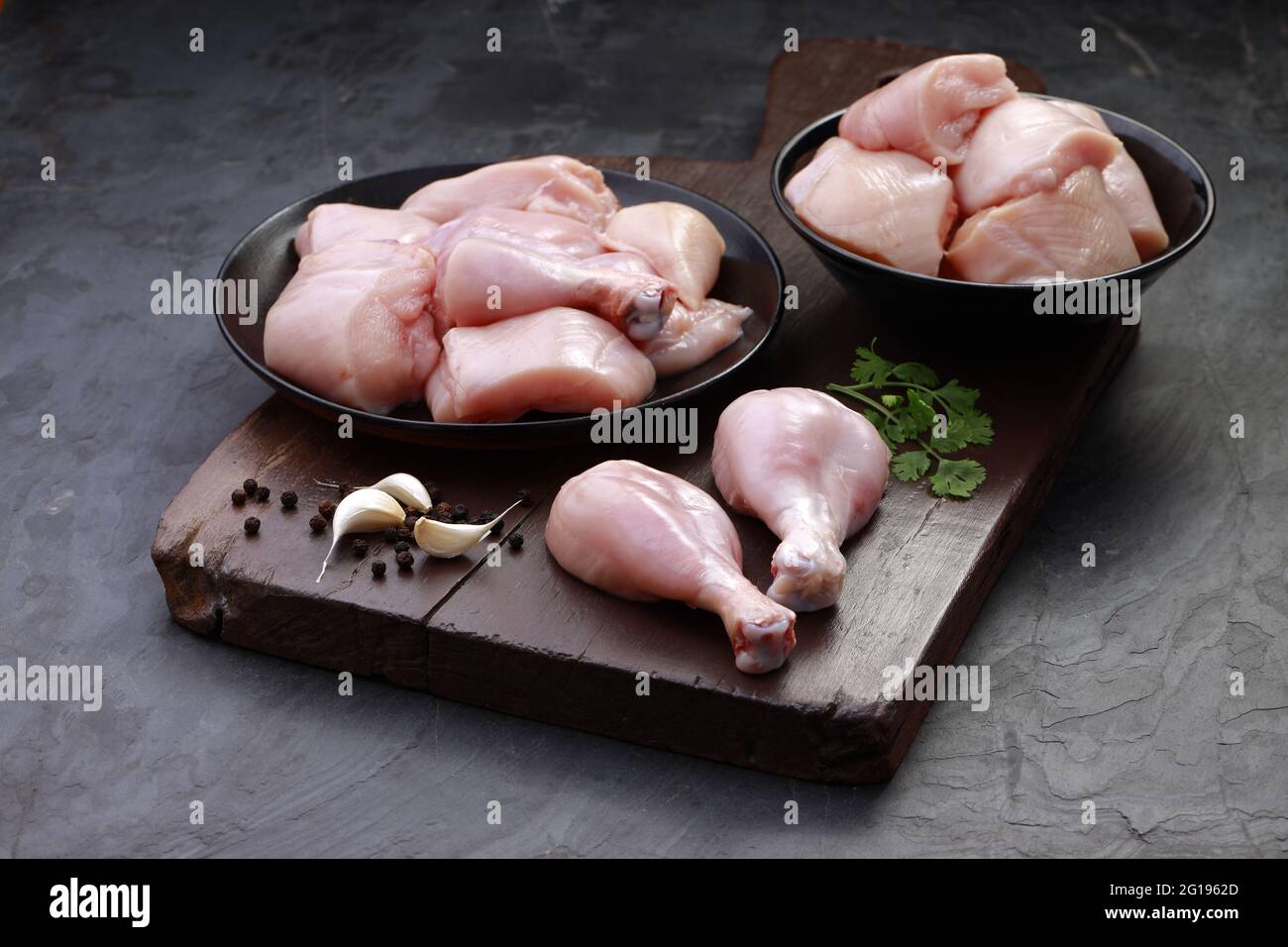 Ensemble de coupes de poulet cru, combinaison de coupes de curry de poulet sans peau, de coupes de poulet et de deux pilons disposés sur une planche en bois avec des feuilles de coriandre Banque D'Images