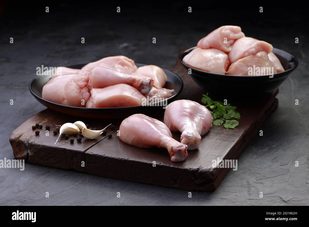 Ensemble de coupes de poulet cru, combinaison de coupes de curry de poulet sans peau, de coupes de poulet et de deux pilons disposés sur une planche en bois avec des feuilles de coriandre Banque D'Images