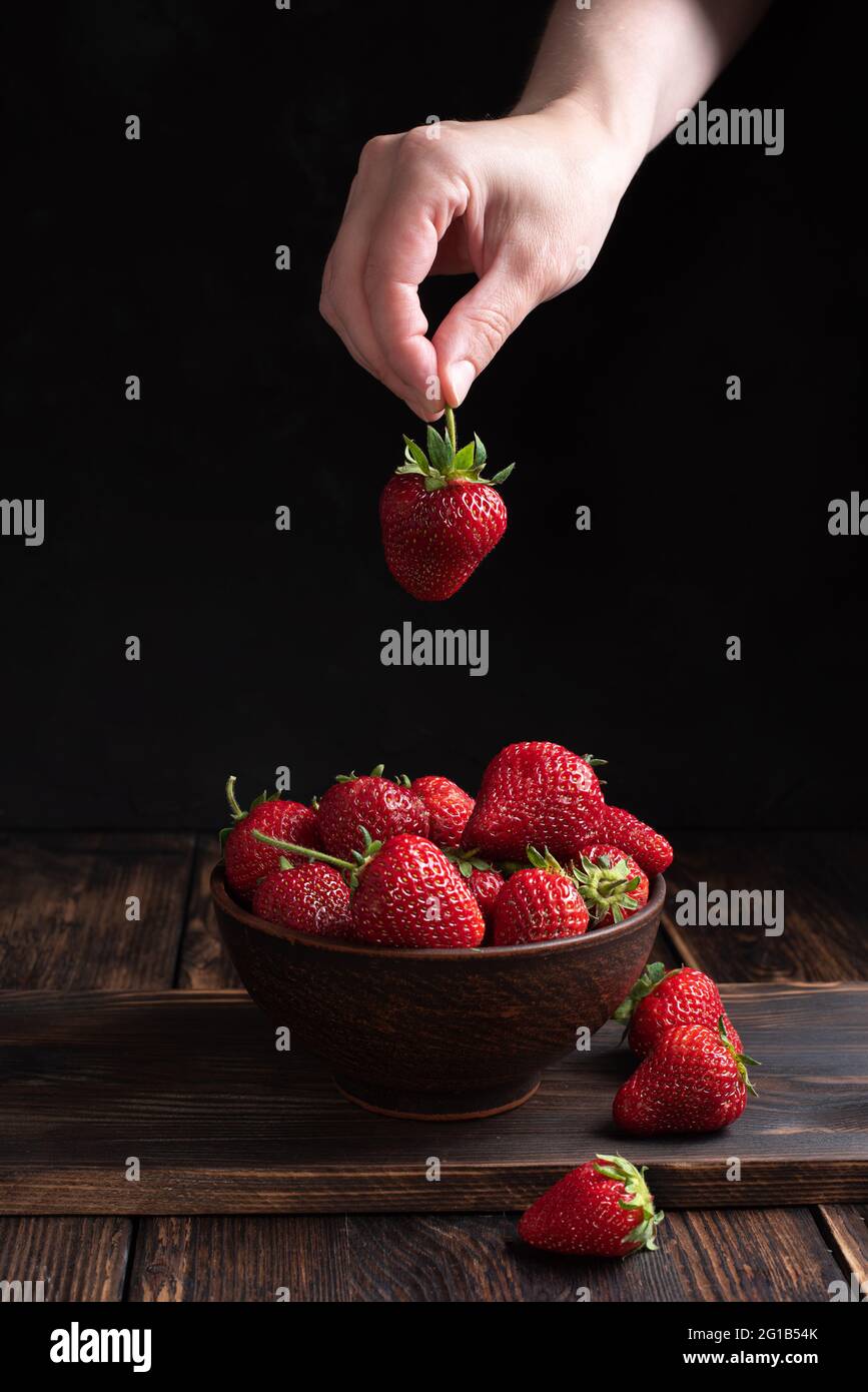 Femme mettant à la main des fraises mûres dans un bol avec des fraises sur fond de bois, style rustique, concept d'été, gros plan. Banque D'Images