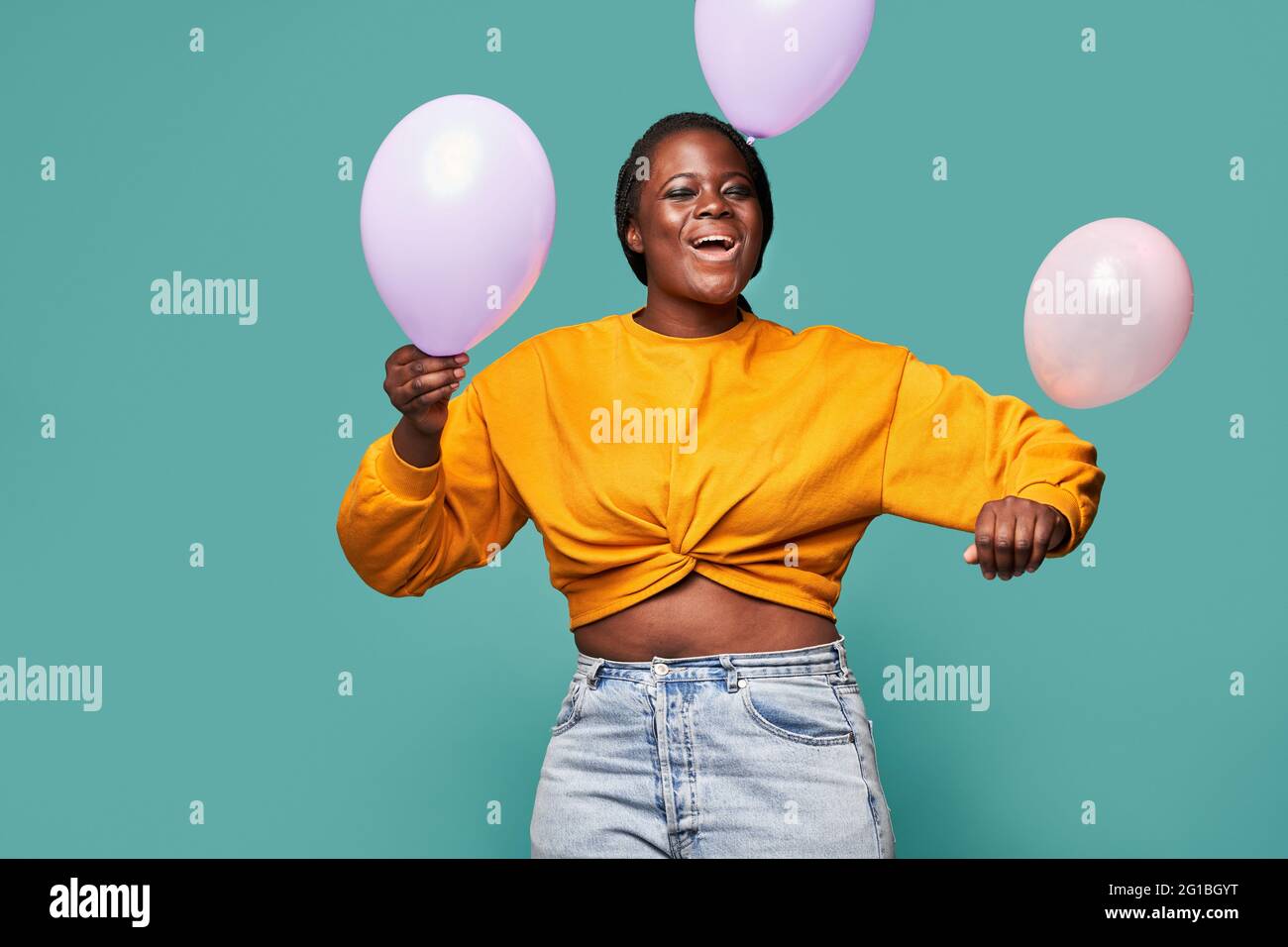Femme afro-américaine excitée en Jean et jaune debout près de ballons tombant sur fond bleu dans le studio Banque D'Images