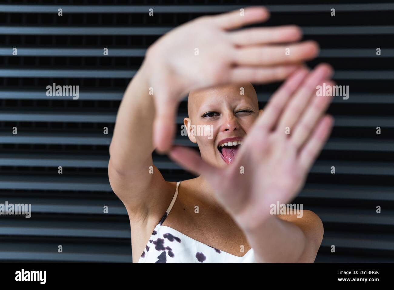 Ravie jeune femme sans cheveux avec alopécie sourire tout en montrant le geste de caméra contre le mur de jalousie dans la rue Banque D'Images
