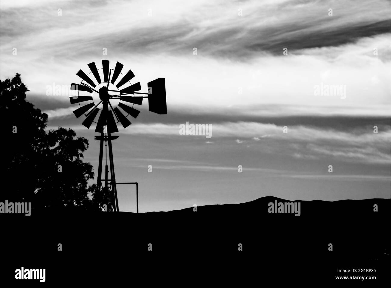 Une silhouette riche, sombre, noire et blanche d'un moulin à vent vintage contre un ciel couvert et nuageux, tout comme le crépuscule prend place dans l'ancien ouest américain. Banque D'Images