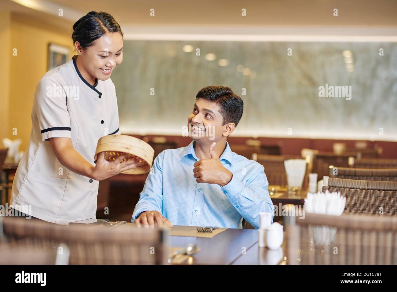 Serveuse souriante donnant une assiette de service avec de délicieux baozi, des petits pains chinois à la viande cuits à la vapeur au client assis à la table et montrant le pouce vers le haut Banque D'Images