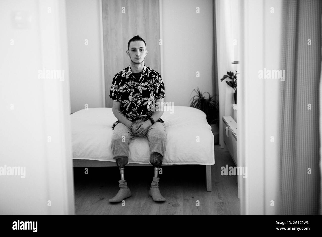 Portrait noir et blanc du jeune homme handicapé assis sur le lit à l'intérieur à la maison, concept prothétique de jambe. Banque D'Images