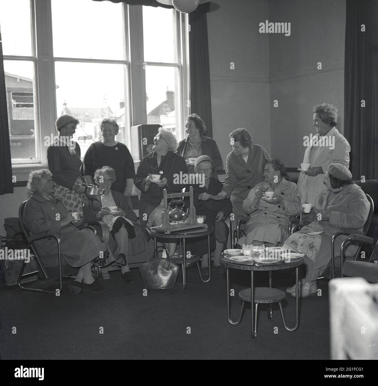 Années 1960, historique, à l'intérieur d'une salle de séjour dans une maison de soins, un groupe de femmes âgées assis dans des chaises autour d'une théière en métal, sur un stand en bois, enfermé « The Singing Kettle Club », étant servi thé et biscuits par le personnel féminin, Écosse, Royaume-Uni. Banque D'Images