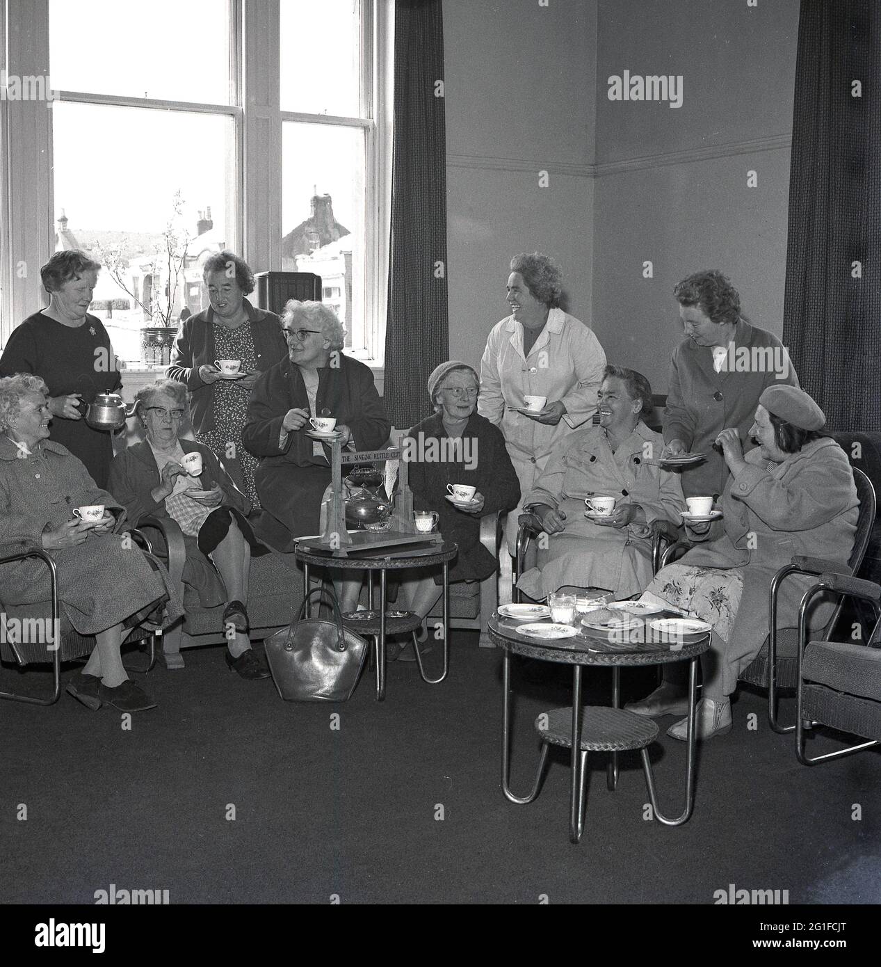 Années 1960, historique, à l'intérieur d'une salle de séjour dans une maison de soins ou de soins infirmiers, un groupe de femmes âgées ayant du thé et des biscuits, assis dans des chaises autour d'une théière plaquée en métal, sur un stand en bois, encrit « The Singing Kettle Club », Fife, Écosse, Royaume-Uni. Banque D'Images