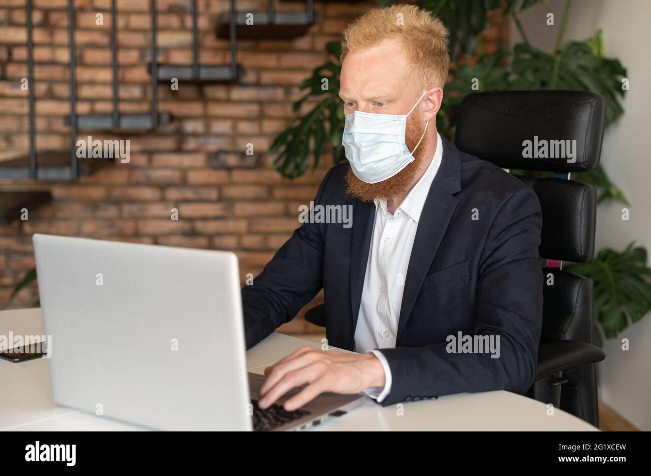 Homme d'affaires sérieux aux cheveux rouges en costume élégant décontracté portant un masque de protection sur un ordinateur portable assis dans un bureau moderne, un employé prend des mesures de protection contre les maladies virales Banque D'Images