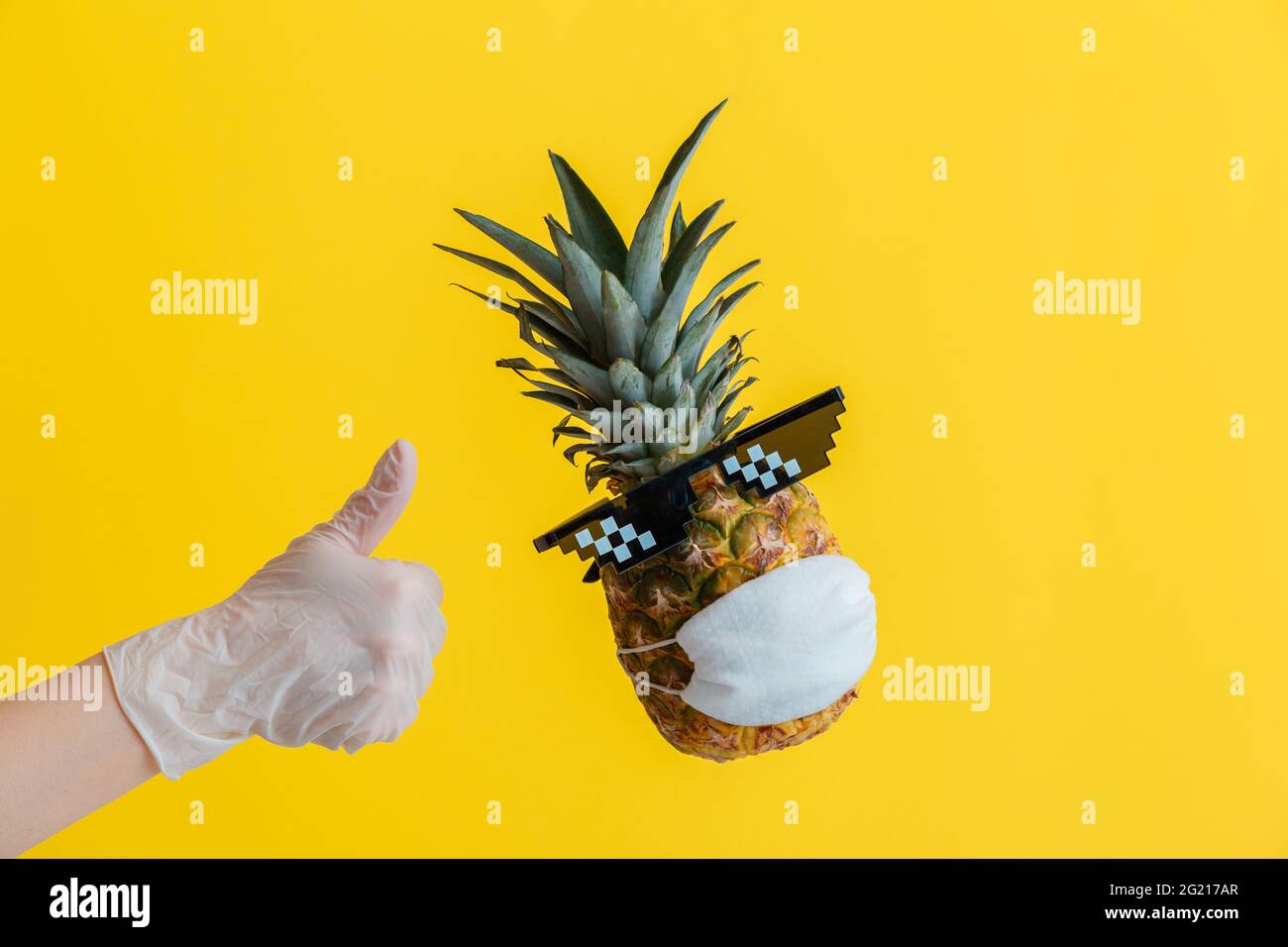 La main dans le gant montre une affiche de pouce vers le haut. Lévitant l'ananas avec le visage drôle portant des lunettes et un masque médical de protection. Concept de coronavirus de voyage Banque D'Images