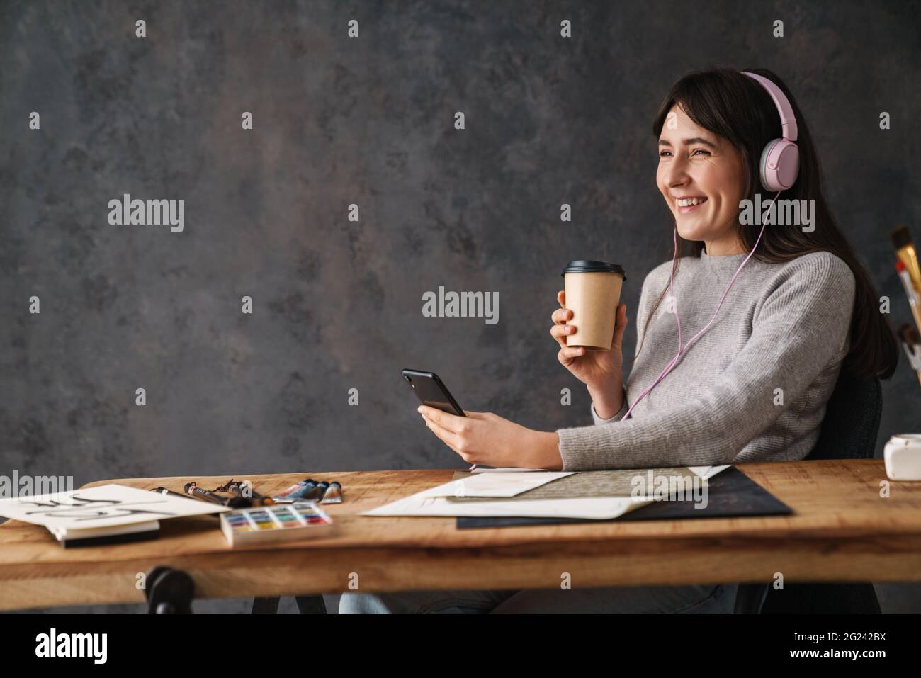 Souriant calligraphe fille dans le casque à l'aide d'un téléphone portable et boire du café tout en travaillant à l'intérieur Banque D'Images