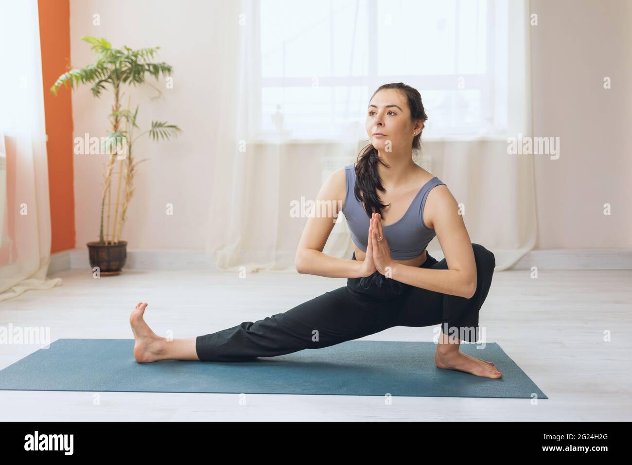 Une femme en pantalon de survêtement et un haut de récolte pratiquant le yoga effectue un exercice de skandasana avec namaste sur un tapis dans le studio. Banque D'Images