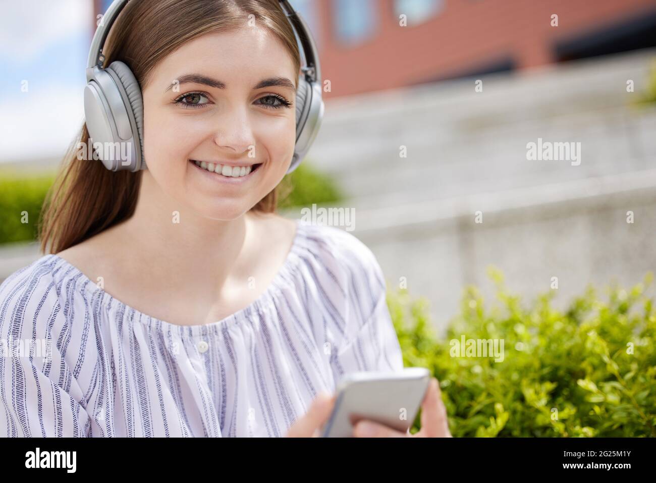 Portrait de la jeune femme étudiante souriante sur le campus de l'université diffuse de la musique du téléphone mobile au casque sans fil Banque D'Images
