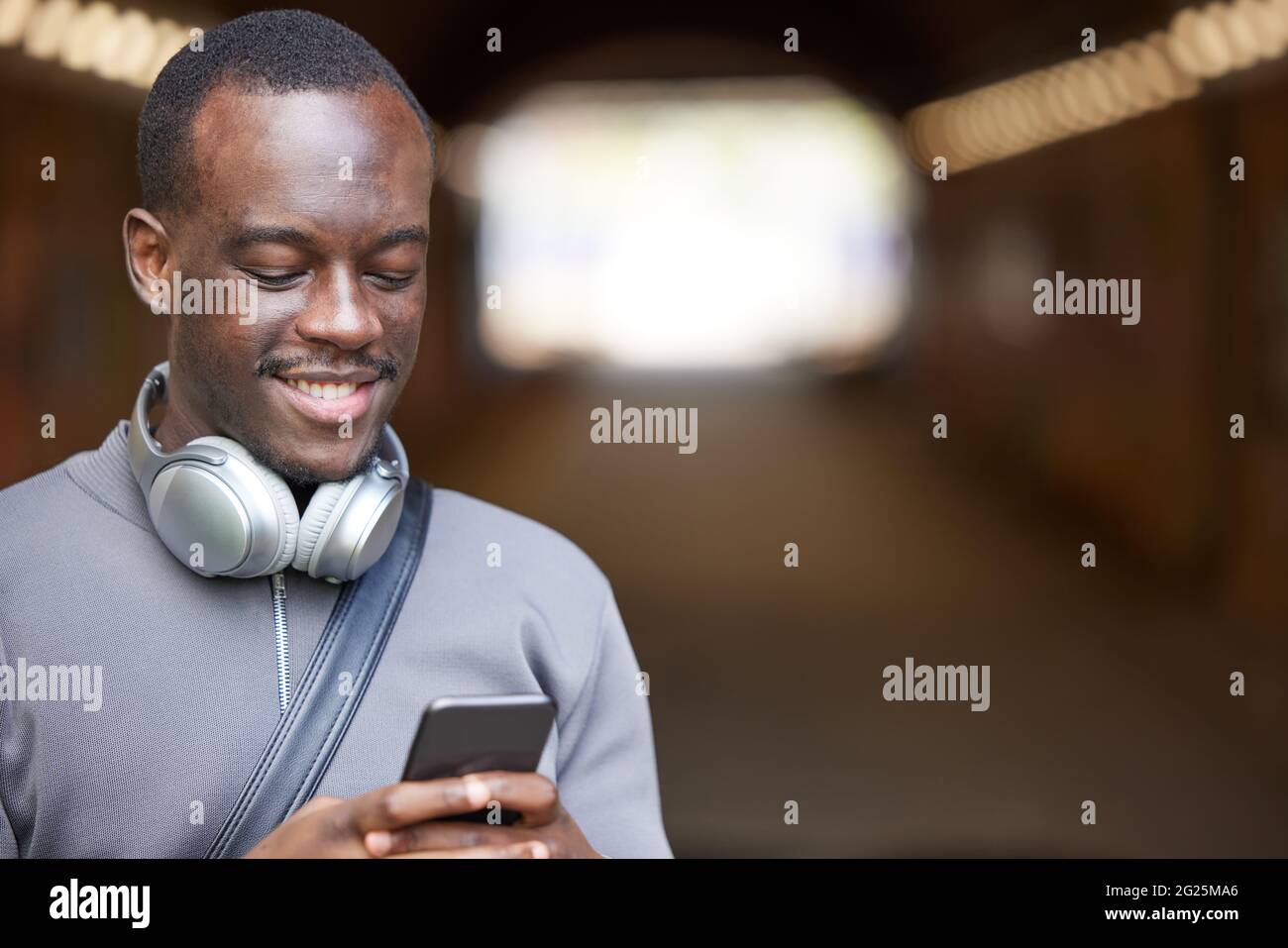 Le jeune homme souriant en milieu urbain vérifie les médias sociaux et les messages sur le téléphone portable tout en portant des écouteurs sans fil Banque D'Images