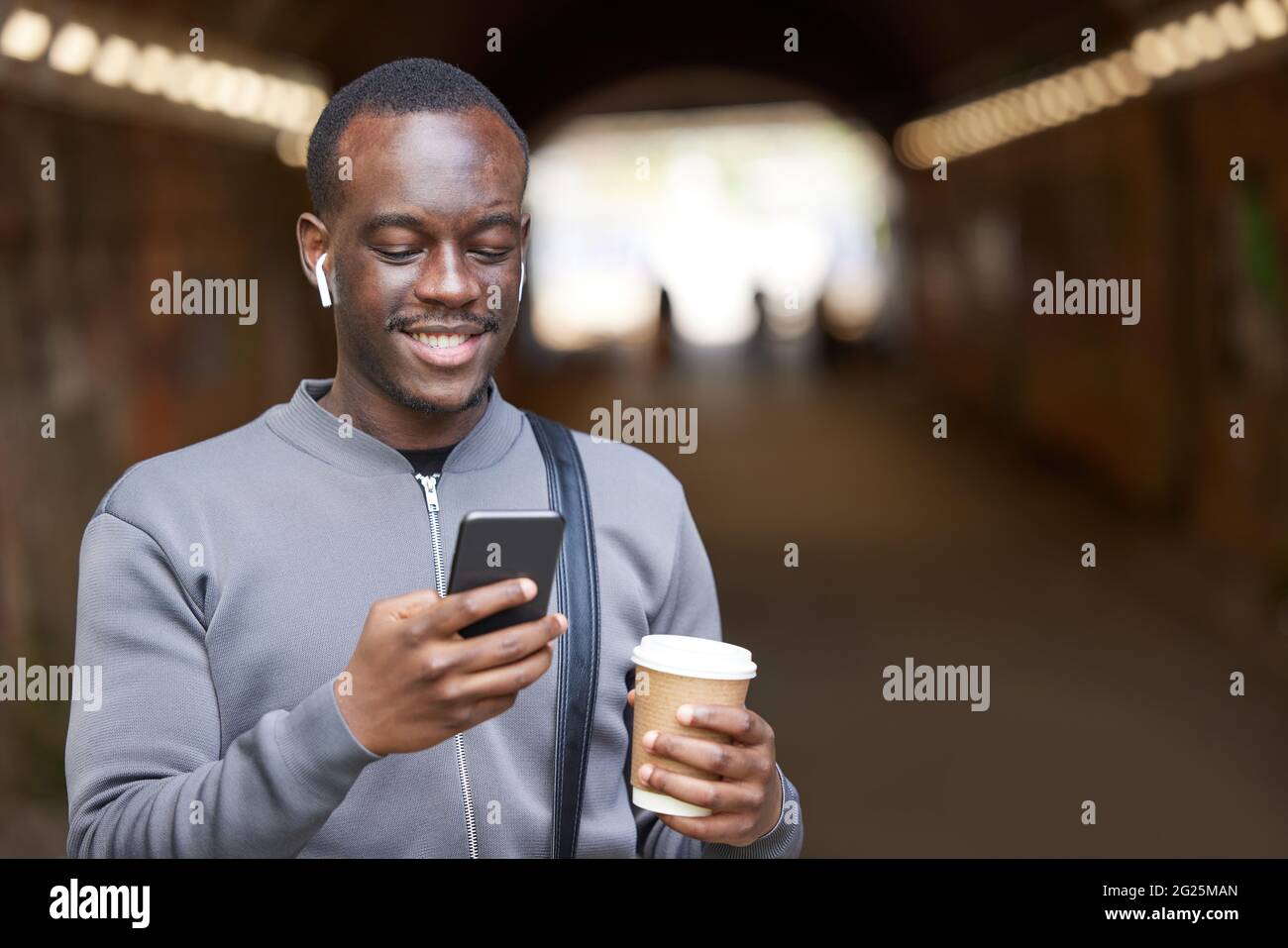 Le jeune homme souriant en milieu urbain vérifie les médias sociaux et les messages sur le téléphone portable tout en portant des écouteurs sans fil Banque D'Images