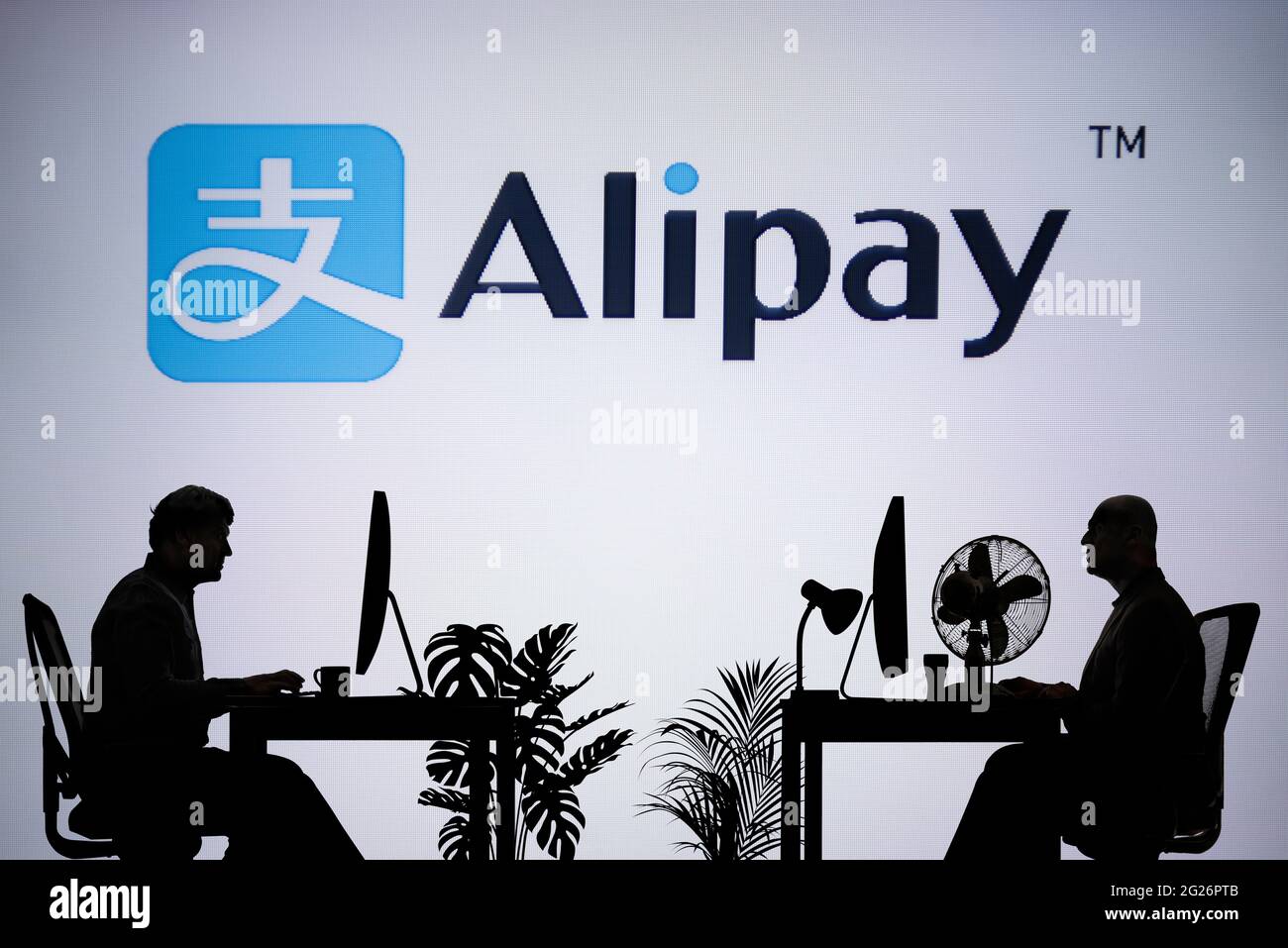 Le logo Alipay est visible sur un écran LED en arrière-plan tandis que deux personnes silhoueteuses travaillent dans un environnement de bureau (usage éditorial uniquement) Banque D'Images