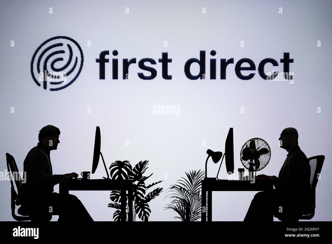 Le logo First Direct est visible sur un écran LED en arrière-plan tandis que deux personnes silhouetées travaillent dans un environnement de bureau (usage éditorial uniquement) Banque D'Images
